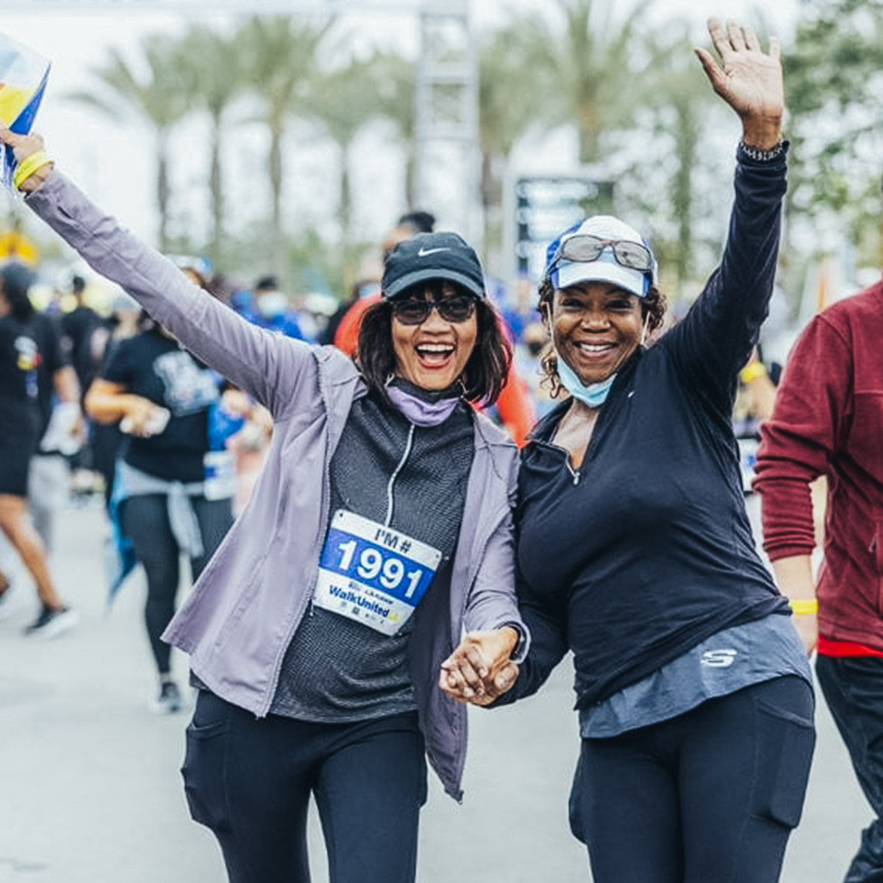 Two women at United Way's 2021 Walk United LA marathon smile and wave at the camera together as people run behind them.