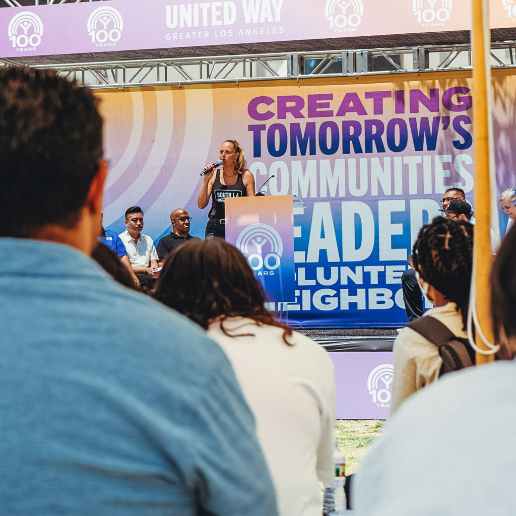 A woman speaks on stage at a podium. Behind her is a massive United Way of Greater L.A. banner that says "Creating tomorrow's communities, leaders, volunteers, neighbors."