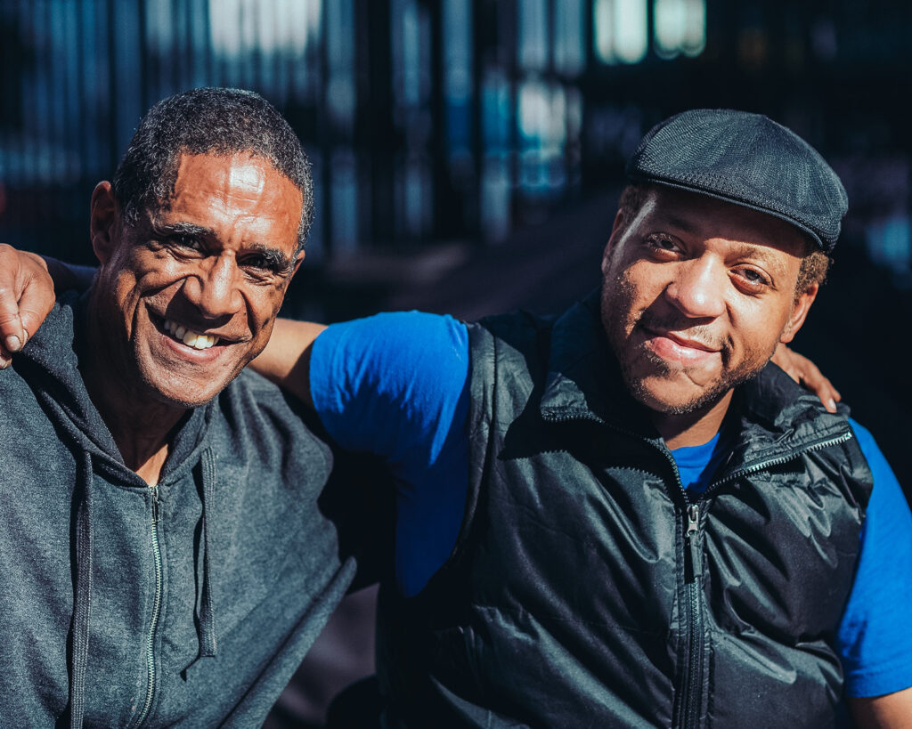 Two men smile at the camera with their arms across each other's shoulders.