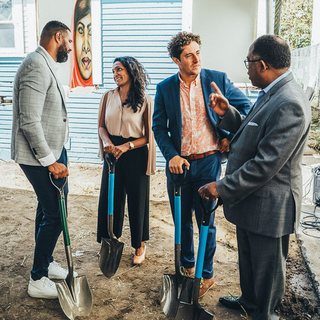 Four people stand in front of a new home development holding shovels and talking.