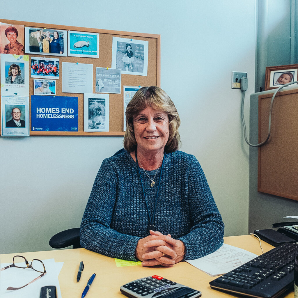 A woman sits at a desk and smiles at the camera. On a cork board behind her there is an Everyone In LA sign that says "Homes end homelessness."