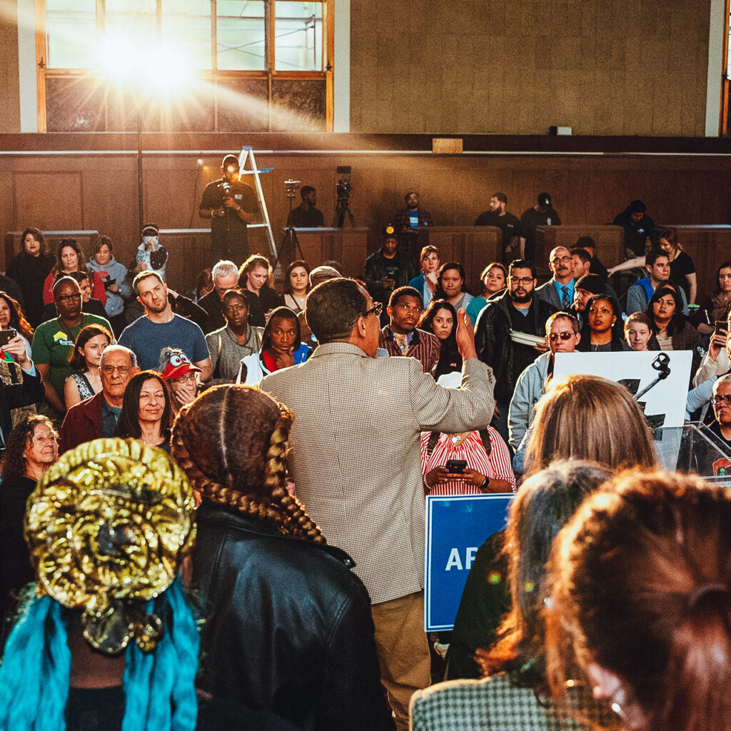 A crowd of people stand around a man speaking into a microphone. Many of the people are wearing shirts that say "Everyone In LA." The room is wide and sunlit.