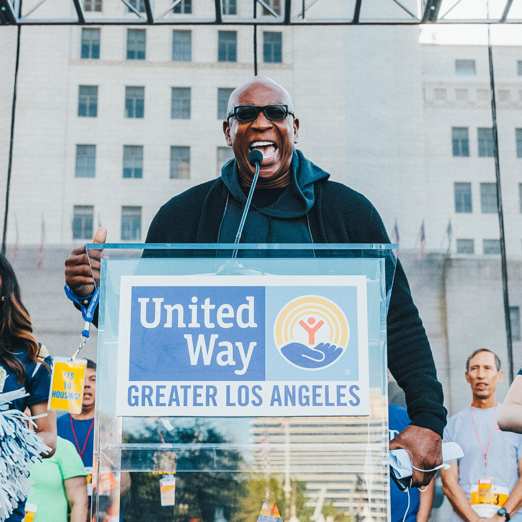 Man of color speaking passionately in front of a podium that says United Way of Greater Los Angeles. Holding a badge that says "Yes to housing".