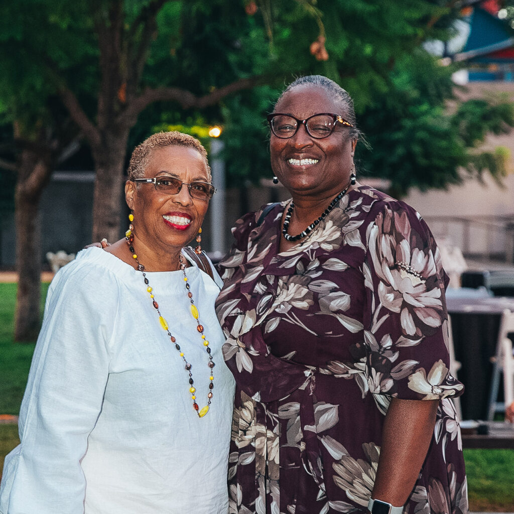 Two women of color stand side by side smiling with their arms around eachother.