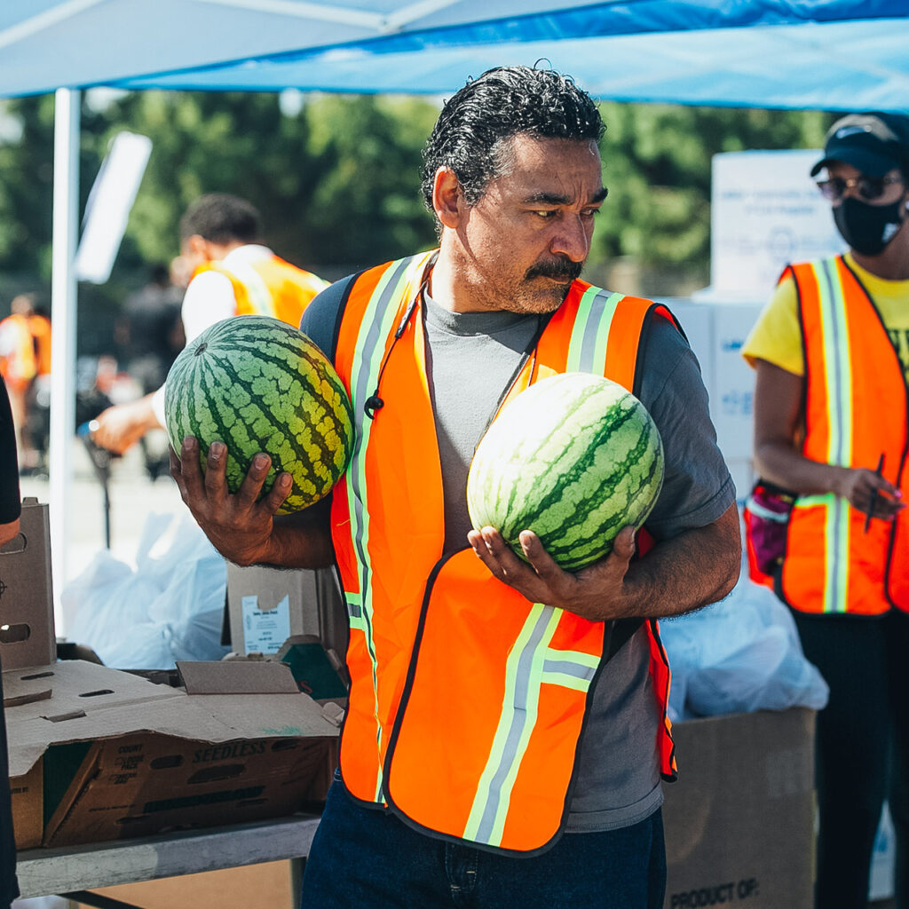 A volunteer in a bright orange safety vest carries two watermelons at a food drive.
