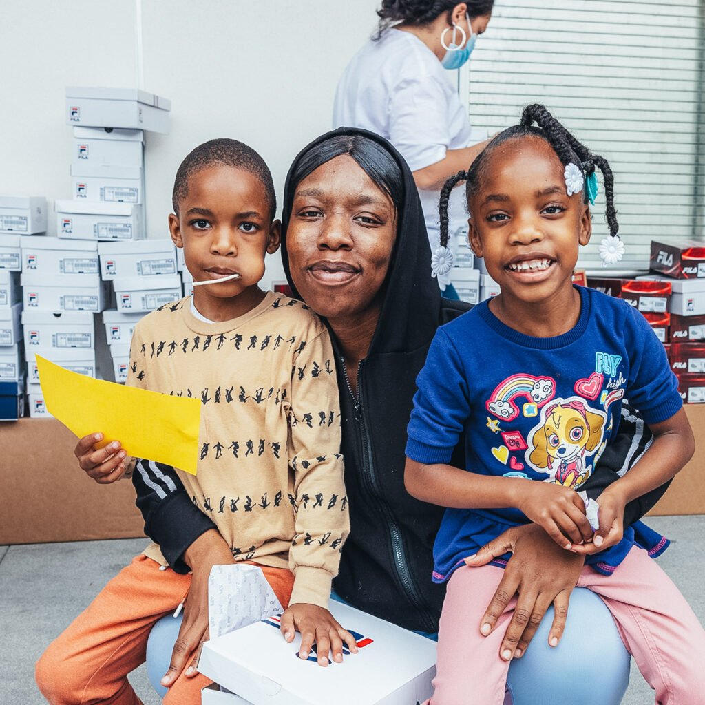 A woman of color sits with two children (one on each knee). The woman and girl on her knee are smiling into the camera while the boy is sucking a lollipop and looking into the camera. There are clothing boxes in the back along with a person in a mask walking behind the family.