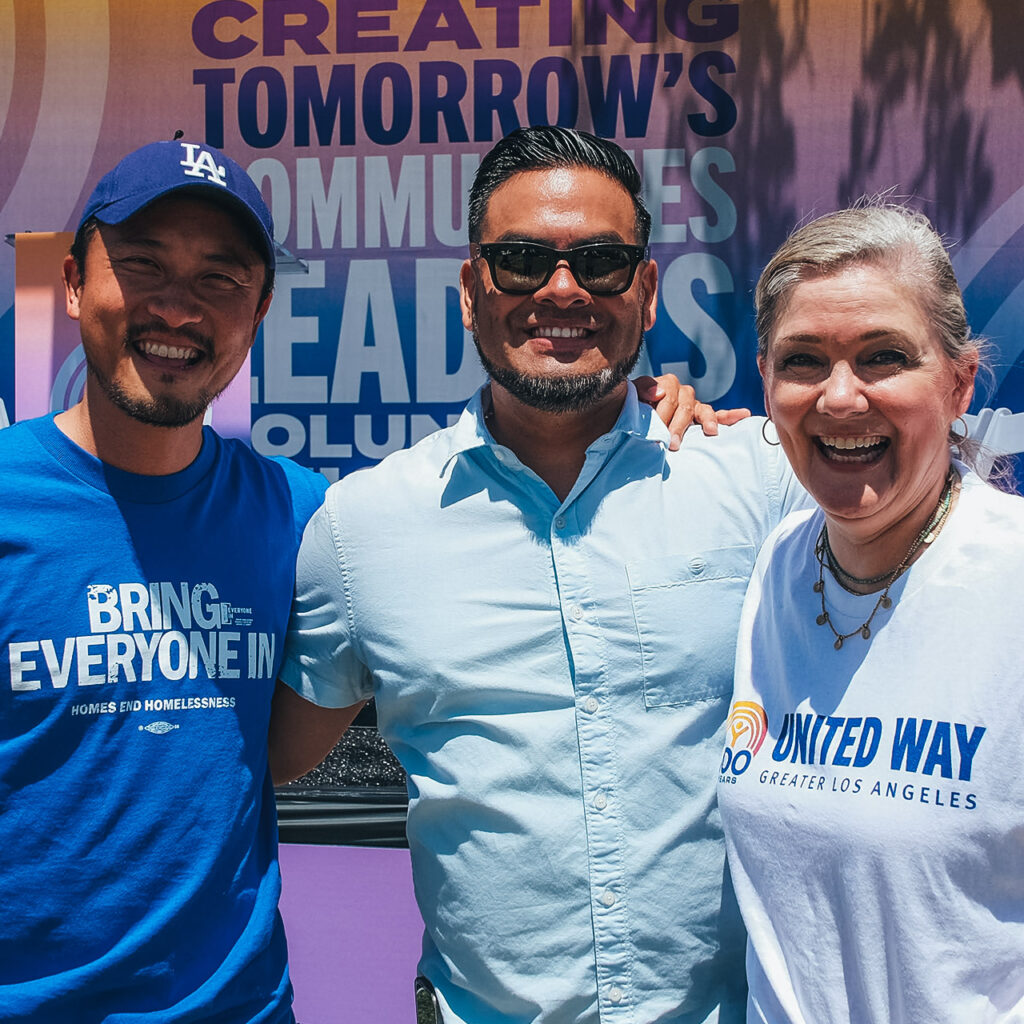 Three people at an outdoor United Way of Greater L.A. event smile at the camera with their arms on each other's shoulders. On the far left, Elise Buik, CEO of United Way of Greater L.A. wears a white United Way shirt. On the far right, a volunteer wears an Everyone In shirt that says Bring Everyone In. In the center, a man wears a pale button down shirt.