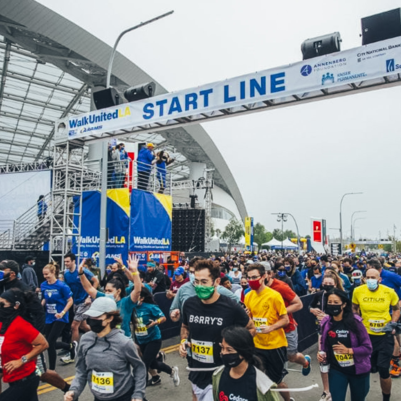 A large crowd of Walk United LA participants jog under a banner that says "Start Line." It's a cloudy day and SoFi Stadium is in the background.