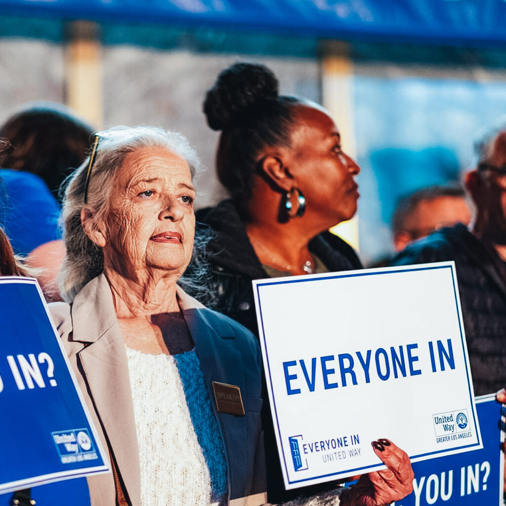 Two women stand near each other holding signs that say Everyone In