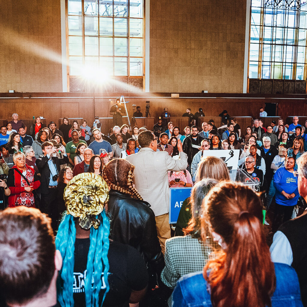 A crowd of people stand around a man speaking into a microphone. Many of the people are wearing shirts that say "Everyone In LA." The room is wide and sunlit.