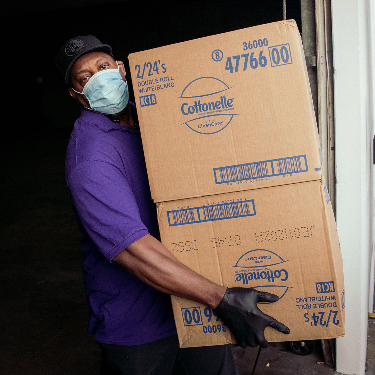 Man wearing purple shirt and mask carries two cardboard boxes.