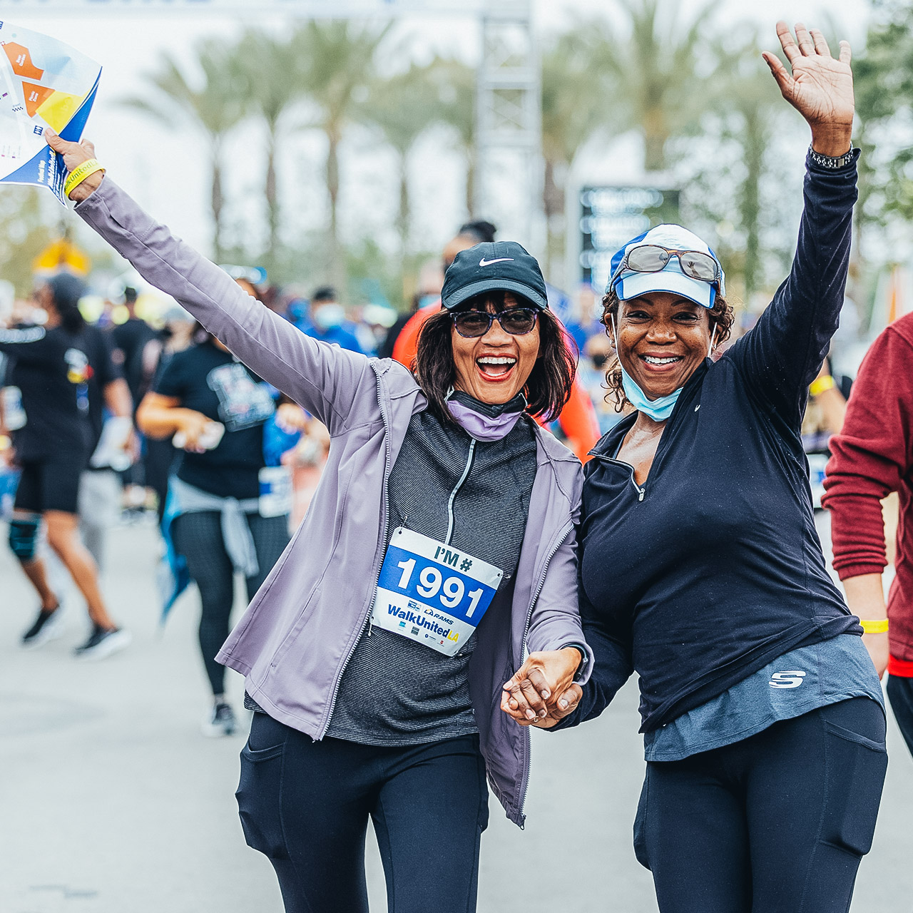 Two women at United Way's 2021 Walk United LA marathon smile and wave at the camera together as people run behind them.